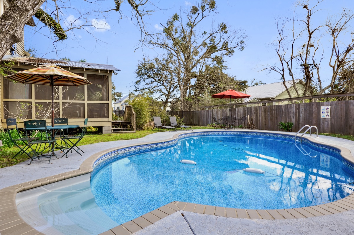 The semi-private saltwater pool is surrounded by a spacious deck. Two umbrellas provide shade over seating areas with metal chairs and a table. Lush greenery and trees enhance the tranquil setting, while a fence encloses the area for privacy.