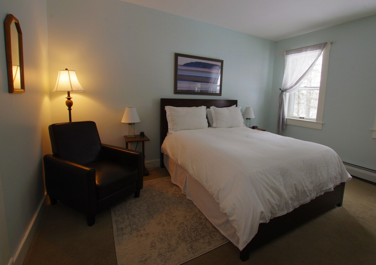 A soothing room features a spacious bed covered in white linens, flanked by two bedside lamps. A comfortable chair is positioned in one corner, and a framed landscape artwork adorns the wall above the bed. Natural light filters in through a window with light curtains.