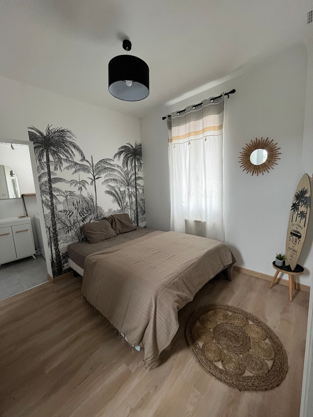 A bedroom featuring a spacious bed dressed in neutral linens. A tropical palm tree mural decorates one wall, while natural light filters through a window dressed with sheer curtains. A round rug is placed on a light wood floor, and a mirror and surfboard add decorative elements.
