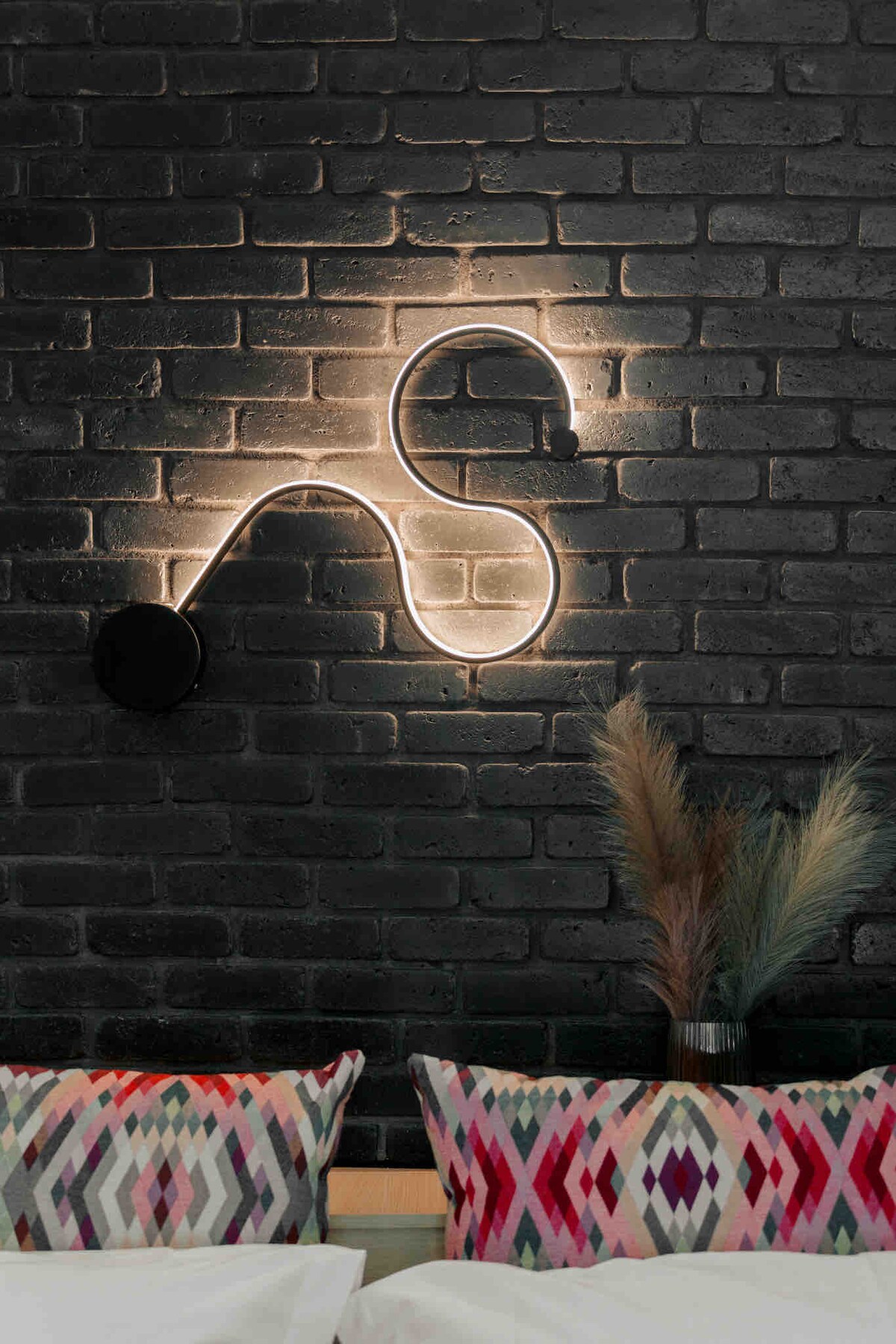 A modern light fixture is mounted on a textured black brick wall, creating a soft illumination with its curved design. Colorful patterned cushions are positioned on a bed, adding a touch of vibrancy to the serene setting.