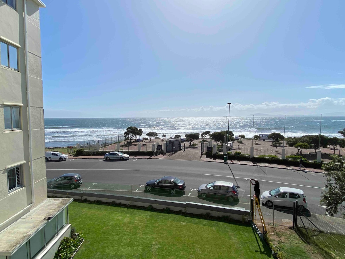 An expansive view of the ocean can be seen from the apartment's window. The bright blue water gently meets the shore, reflecting sunlight. A manicured lawn is visible in the foreground, with a roadway and parked cars beyond, enhancing the coastal setting.