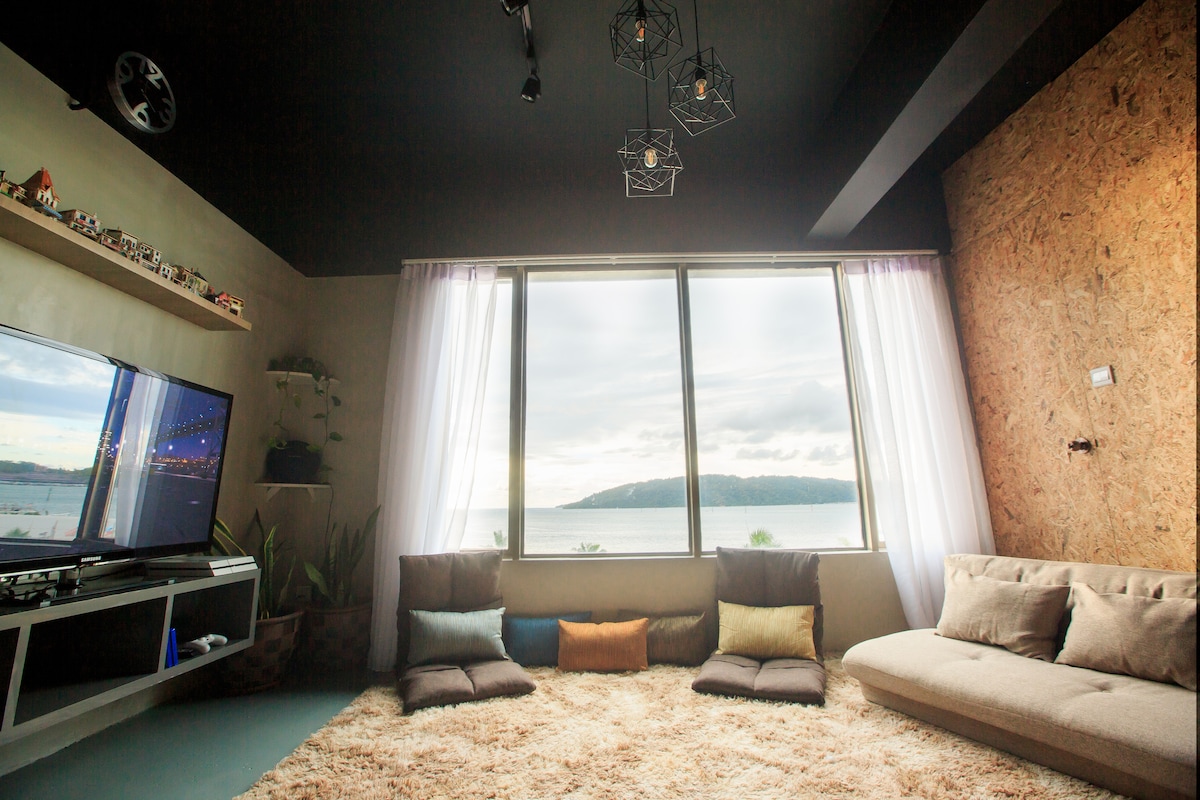 A common area features large windows framing a scenic sea view. Soft furniture, including a sectional couch and floor cushions, is arranged on a plush rug. Natural light brightens the space, complemented by decorative elements and overhead lighting.