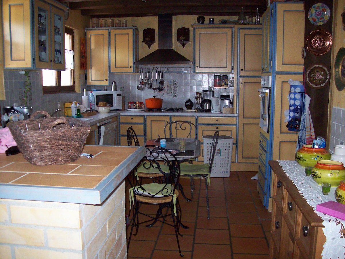 A country-style kitchen features light blue cabinetry with yellow accents and traditional ceramic plates on the walls. A wooden dining table with four chairs is positioned near a brick island, while various kitchen appliances are visible on the countertops.