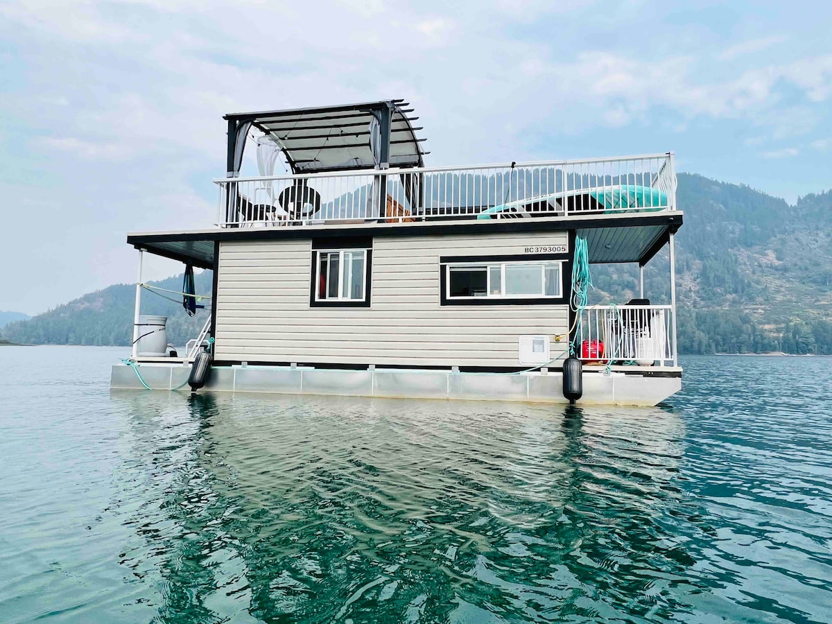 A spacious cabin is anchored on clear waters, featuring a deck with a shaded seating area. The cabin is designed with large windows, providing views of the surrounding nature. The tranquil lake reflects the cabin, surrounded by gentle hills in the background.