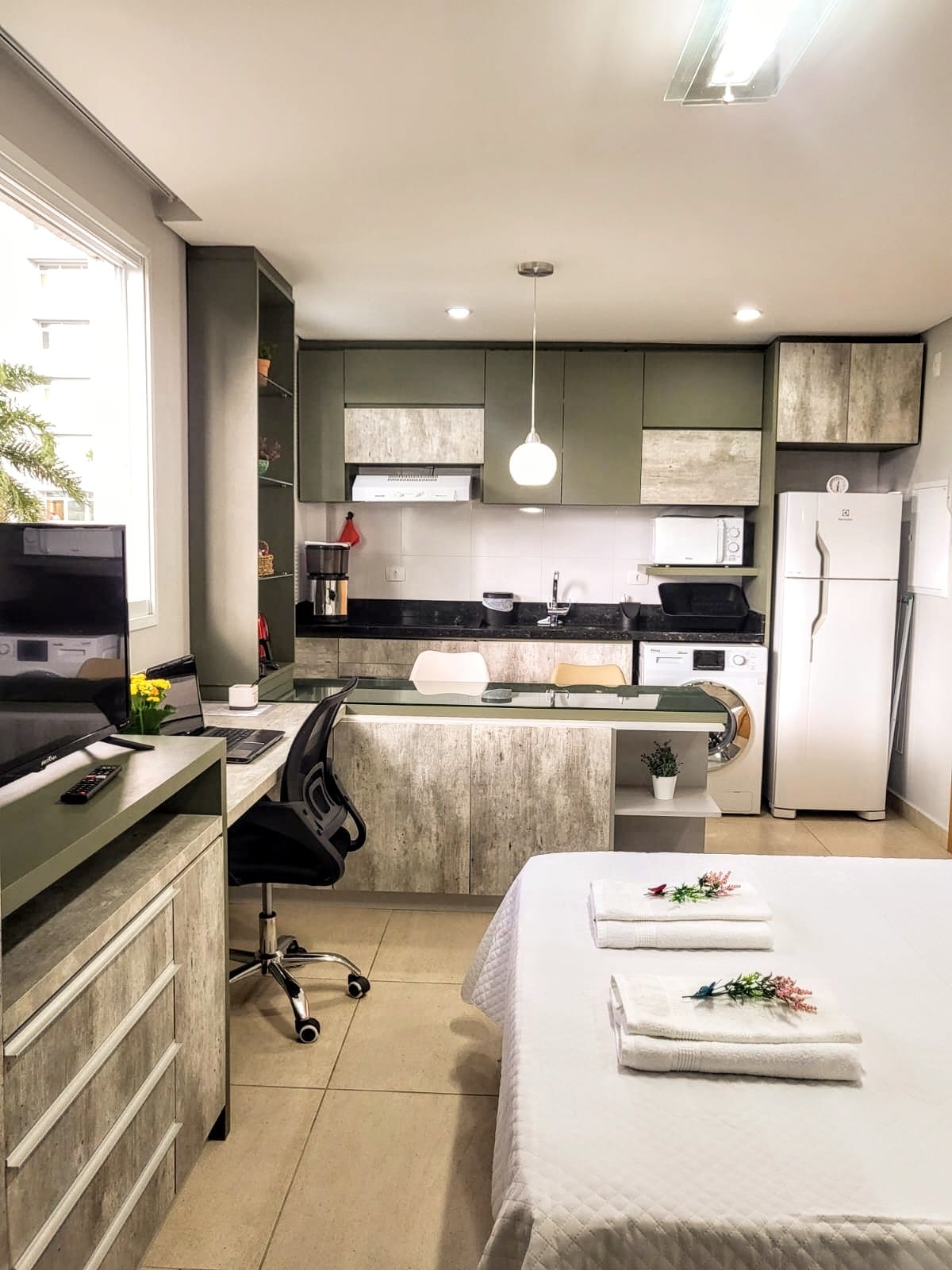 A well-designed studio features modern cabinetry in muted green and wood finishes. A workspace with a desk chair is positioned next to a window. The open kitchen is fully equipped with essential appliances, and a neatly made bed is adorned with folded towels and decorative flowers.