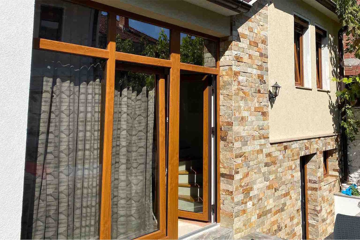 A well-crafted exterior features a combination of natural stone and smooth wall surfaces. Large wooden-framed windows allow natural light to enter, showcasing an inviting entrance. A wall lantern adds a touch of charm, enhancing the area's aesthetic appeal.