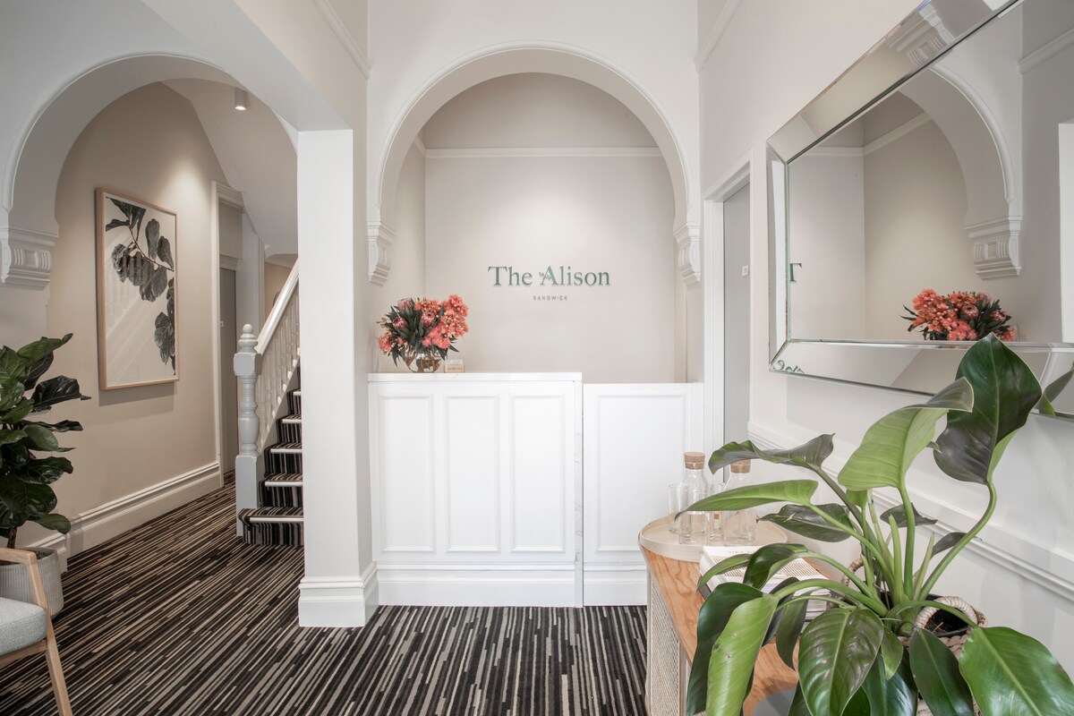 A bright and welcoming entrance area features arched doorways and elegant flooring. Neutral walls are complemented by a large mirror and decorative plants. A staircase is visible in the background, leading to additional rooms. Floral arrangements add a touch of color to the space.