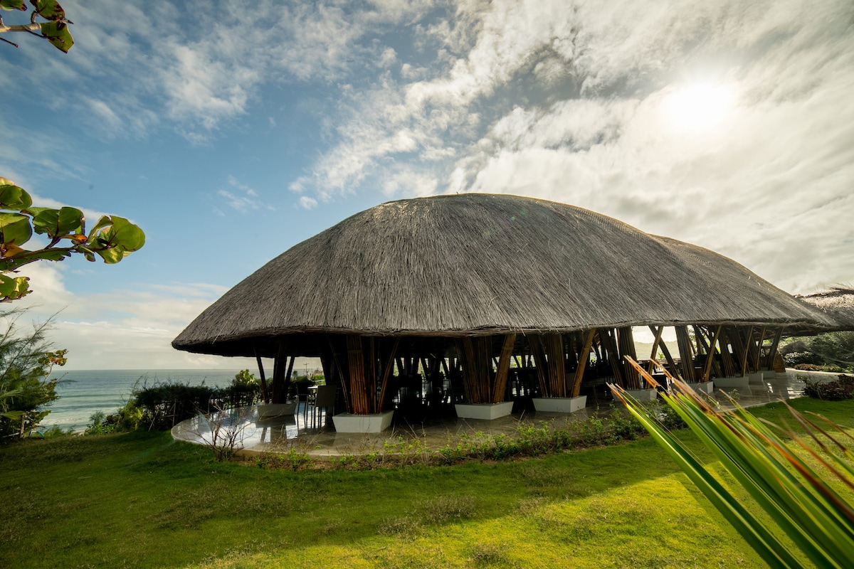 A unique structure with a thatched roof is set against a backdrop of blue sky and ocean. Wooden supports elevate the design, surrounded by lush greenery. The space is designed to harmoniously blend with nature, providing views of the seascape.