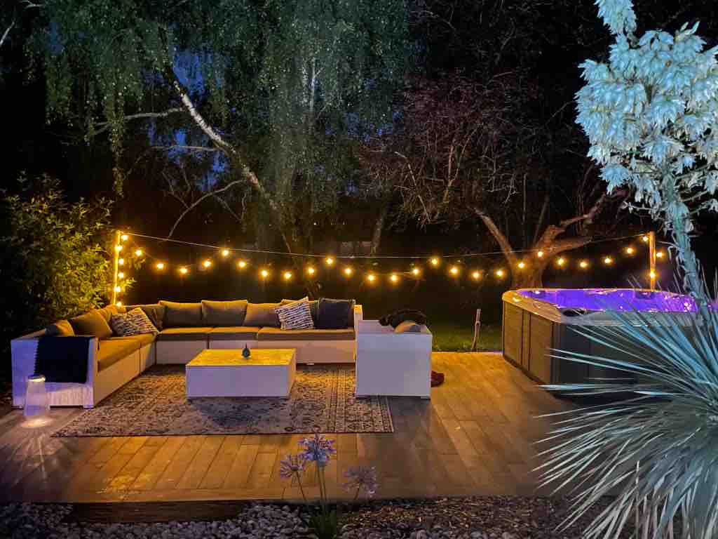 An outdoor lounge area is illuminated by warm string lights, creating a cozy ambiance at night. A spacious seating arrangement faces a central coffee table, complemented by a patterned rug. A jacuzzi sits nearby, surrounded by greenery, enhancing the serene outdoor setting.