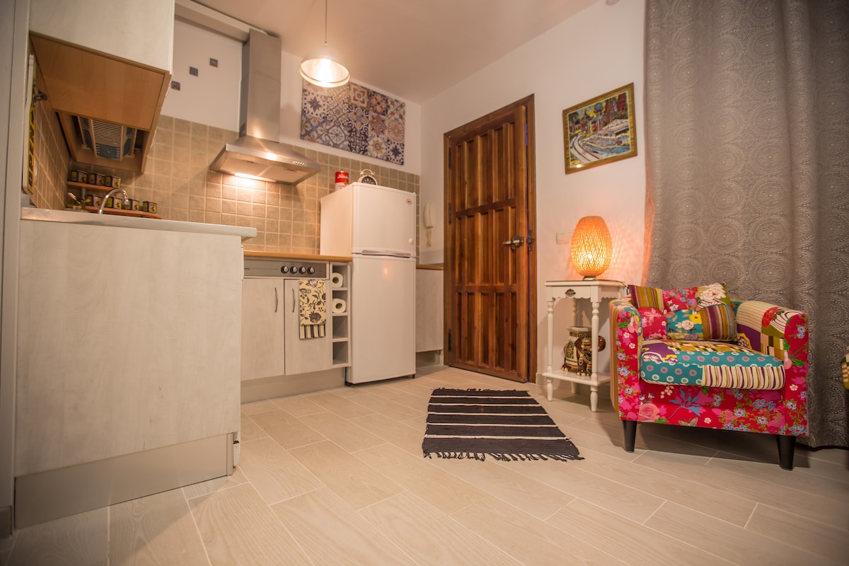 A compact kitchen area offers modern appliances including a stove, refrigerator, and wooden cabinetry. A decorative chair adorned with colorful fabric adds a pop of color. Soft lighting illuminates the space, while a textured curtain drapes elegantly beside the door.