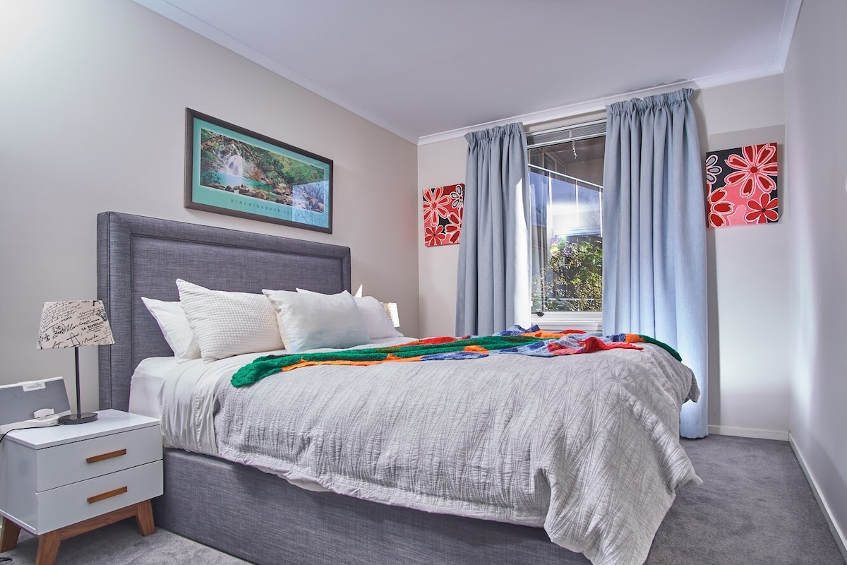 A spacious bedroom features a comfortable bed layered with soft linens and a colorful throw. Light filters through a window adorned with flowing curtains, while framed artwork and vibrant decor accents the walls, contributing to a welcoming atmosphere.