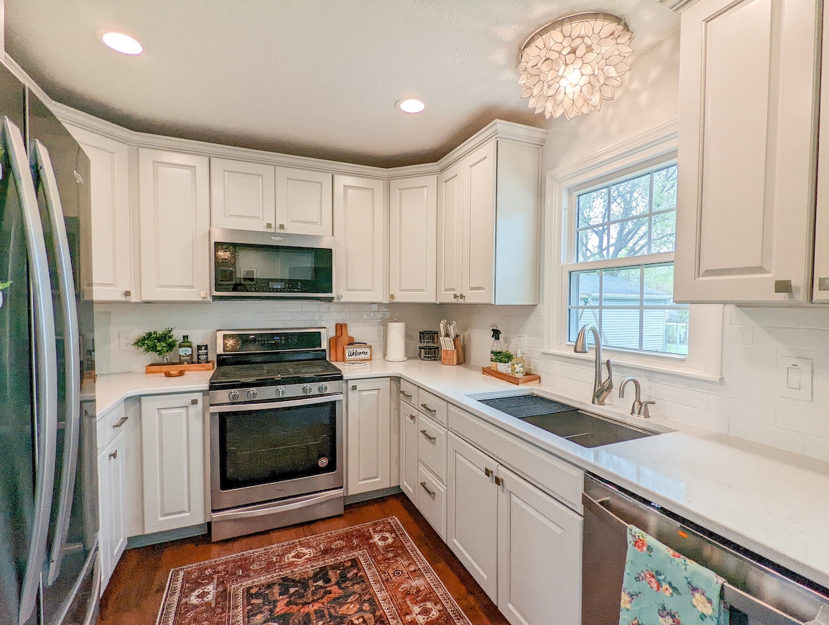 The modern kitchen features light gray cabinetry, quartz countertops, and stainless steel appliances. An extra deep sink is positioned under a window, allowing natural light to illuminate the workspace. A decorative light fixture hangs from the ceiling, adding a stylish touch.