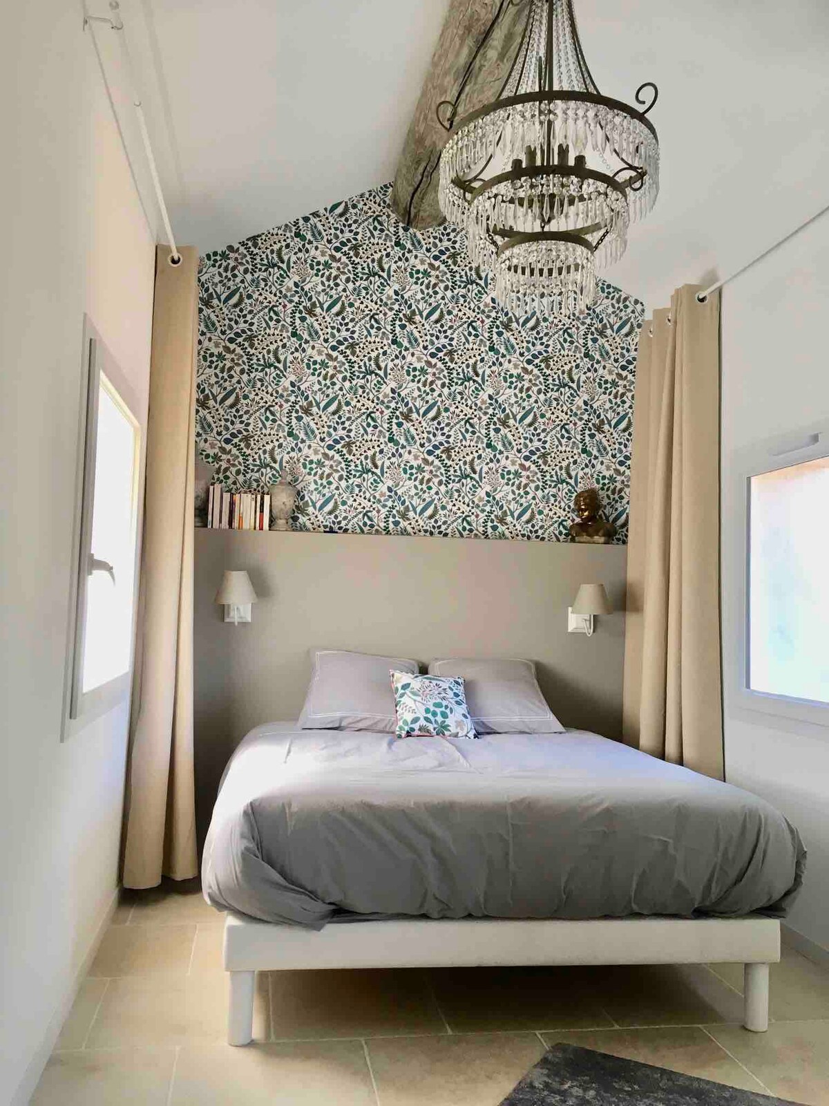 A spacious bedroom features a queen-sized bed draped in soft gray linens. An intricately patterned wallpaper adorns the wall behind the bed. Two windows provide natural light, with lightly colored curtains framing the space. A crystal chandelier hangs above, adding an elegant touch.