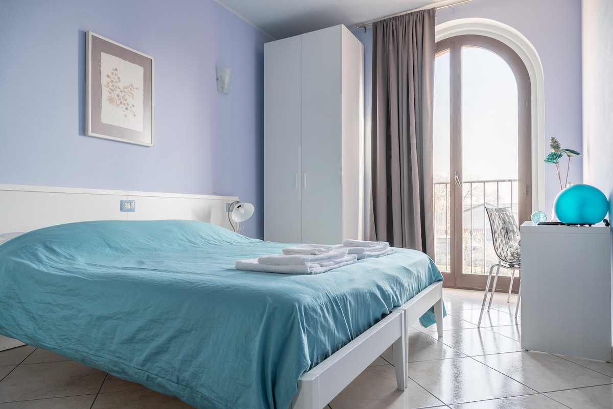 A bright bedroom features a spacious bed covered with a light blue blanket. A tall wardrobe stands against the wall, while a modern chair is placed near the window. Sunlight fills the room through an arched window, complemented by soft gray curtains.
