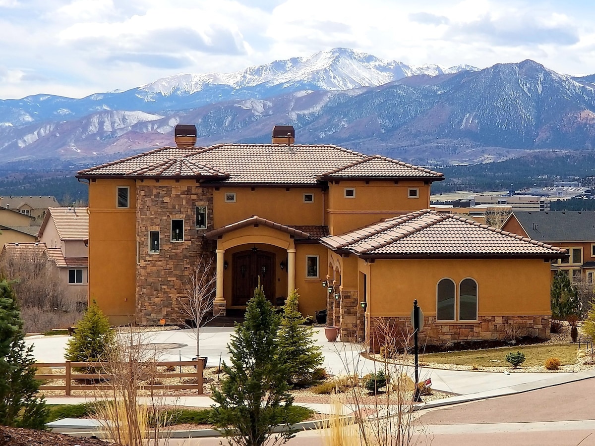 A grand Tuscany-style retreat is showcased against a backdrop of snow-capped mountains. The building features earthy tones and an elegant entrance, surrounded by landscaped greenery. The driveway leads to a circular area with manicured shrubs, highlighting the home's spacious architecture.