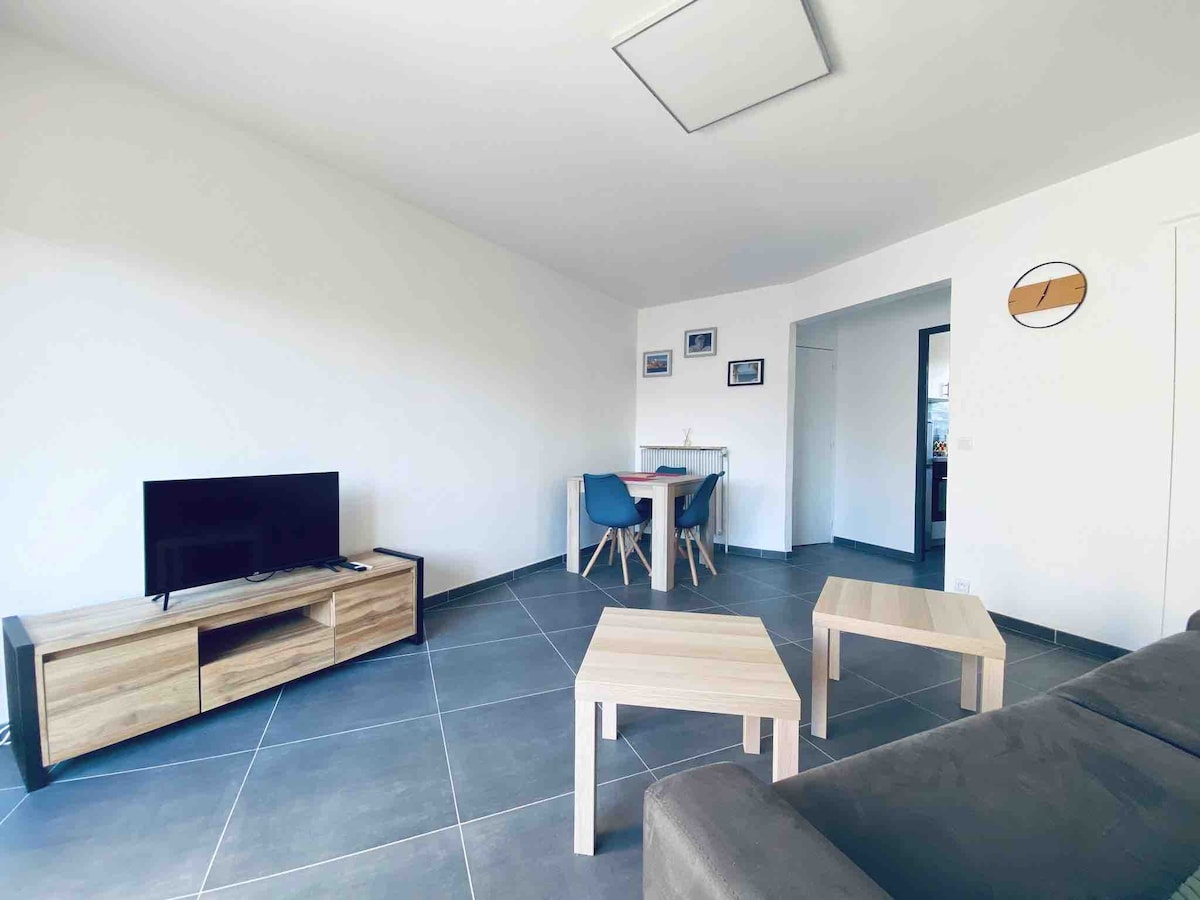 A bright living area features a modern design with a comfortable gray sofa and a low coffee table. A small dining table with blue chairs is positioned nearby, and a television is placed on a wooden media console. Large windows allow natural light to fill the space.