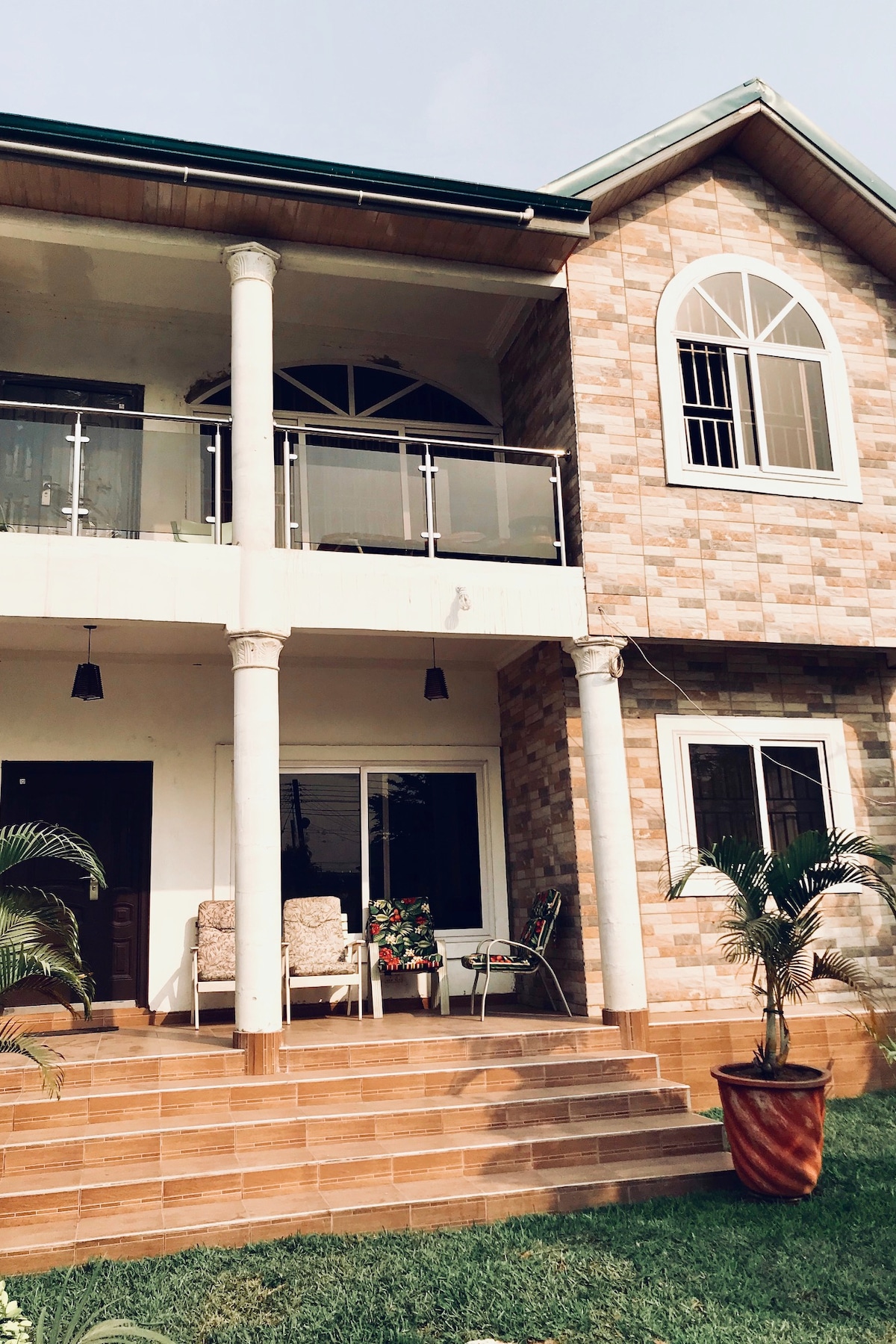 The exterior of a two-story house is featured, showcasing a stone facade and large front steps. A spacious balcony with glass railings provides seating options. Plants are visible near the entrance, enhancing the welcoming appearance of the home.