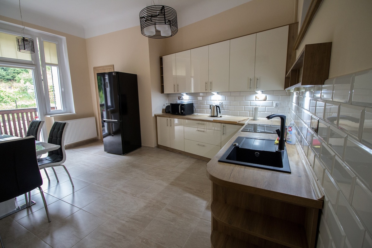 A modern kitchen is displayed with sleek cabinetry and stainless steel appliances, including a refrigerator and kettle. A large window allows natural light to brighten the space. A dining area with black chairs is positioned beside a small wooden table, creating a functional and inviting environment.