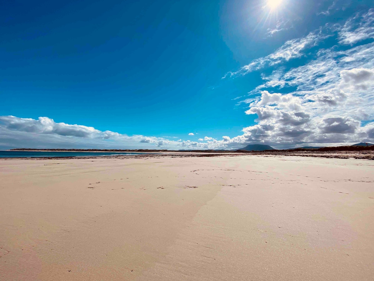 A wide view of a sandy beach unfolds under a bright blue sky, scattered with soft white clouds. The shoreline appears calm and inviting, with gentle textures visible in the sand, highlighting the natural beauty of the coastal landscape.