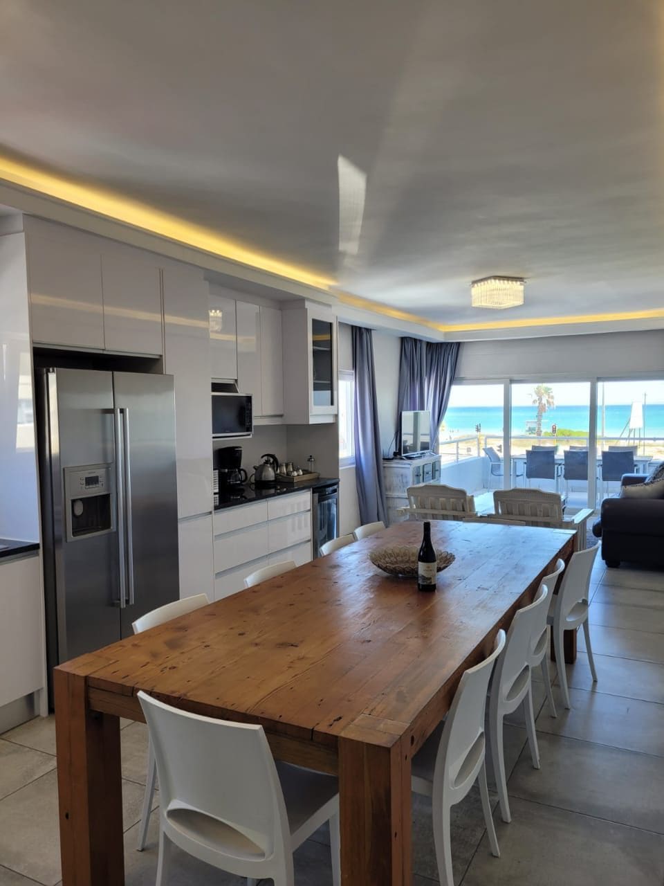 A modern kitchen area features a large wooden dining table surrounded by white chairs. Sleek cabinetry and stainless steel appliances are visible, with a view of the sea beyond large windows, allowing natural light to brighten the space.