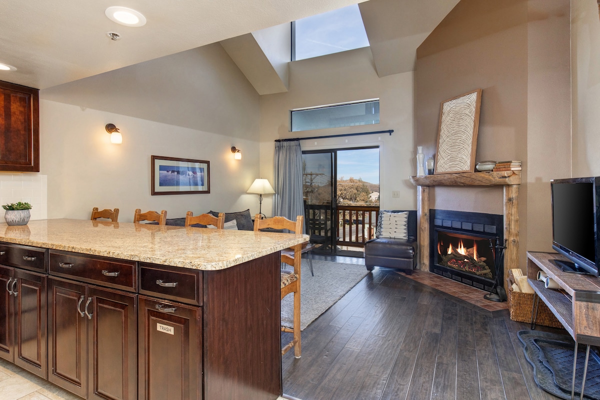 The open-concept living and kitchen area features a spacious island with bar seating, warm wood cabinetry, and a cozy fireplace. Large windows provide natural light and a view of the outdoors, while a sliding door offers access to a small balcony.