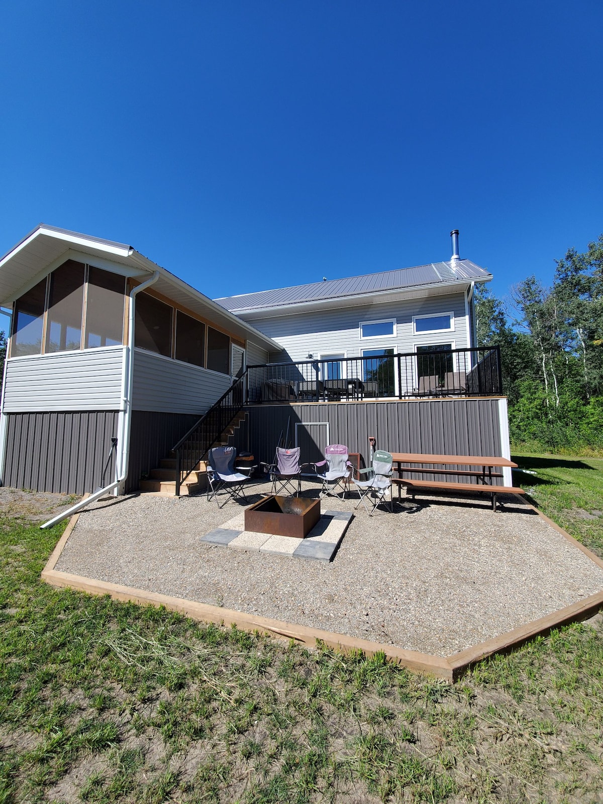 An outdoor seating area features several chairs arranged around a fire pit, set on a gravel surface. The house is visible in the background, showcasing a screened-in porch and large windows. Lush greenery surrounds the space, creating a serene atmosphere.