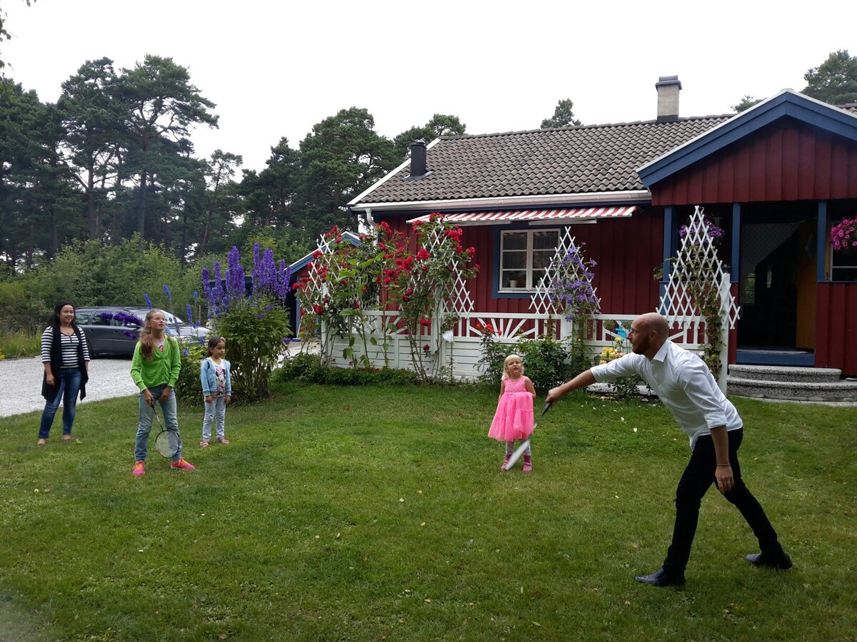A red wooden house is visible, featuring a sloped roof and a welcoming porch. Colorful flowers line the front, with trellises adding charm to the garden. A grassy area is occupied by a group of children and adults, engaged in playful activities.