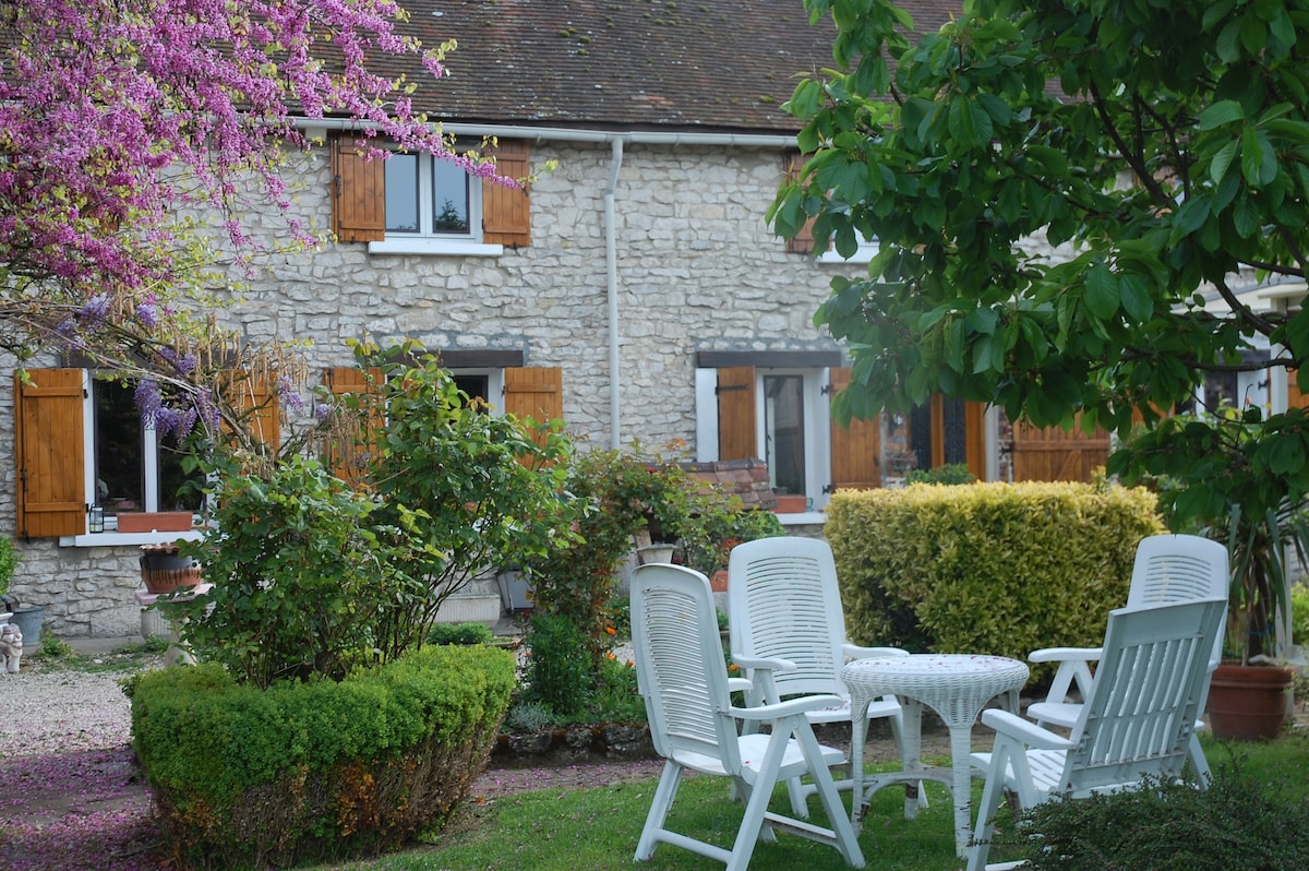 A stone building is framed by flowering trees and greenery, with wooden shutters and a gravel pathway leading to the entrance. A circular garden area is surrounded by neatly arranged white outdoor furniture, offering a peaceful spot for relaxation amid the natural setting.