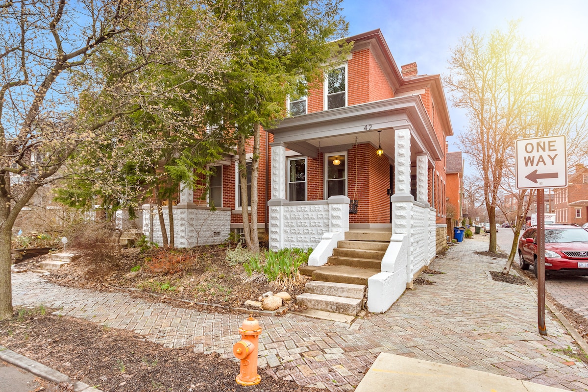 The exterior of the home features a red brick facade with white stone accents and a covered front porch. Steps lead to the entrance, surrounded by flowering plants and trees. A stone-paved walkway is visible alongside the house, enhancing the home's charm.