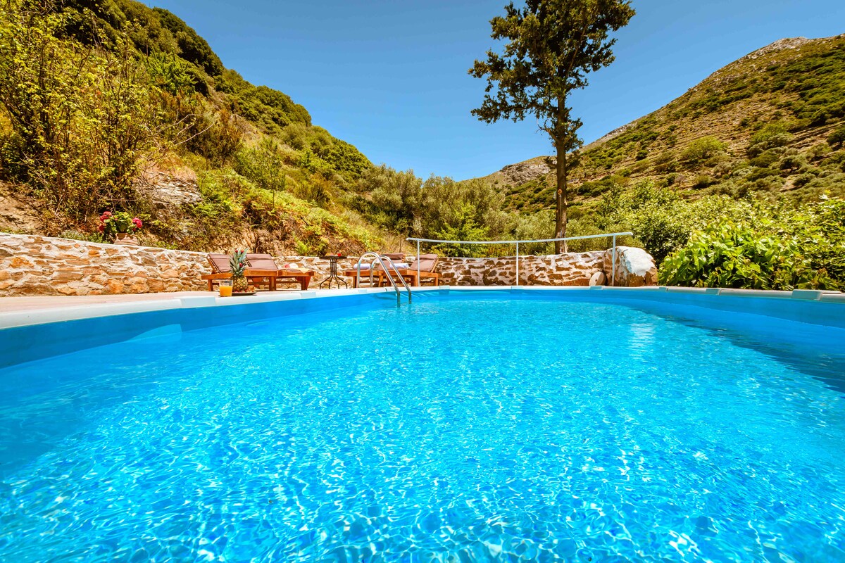 A clear blue swimming pool is surrounded by natural greenery and stone walls. Sun loungers are positioned nearby, providing a relaxing space to enjoy the serene landscape. Rolling hills and trees frame the background, enhancing the tranquil atmosphere.