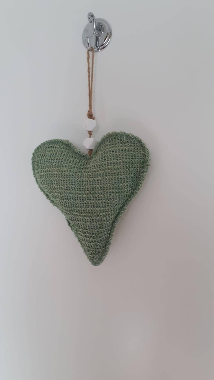 A heart-shaped decoration made from woven material is suspended from a silver hook. The green hue of the heart contrasts against the white wall, and small decorative beads are attached to the hanging cord, adding a subtle detail to the overall design.