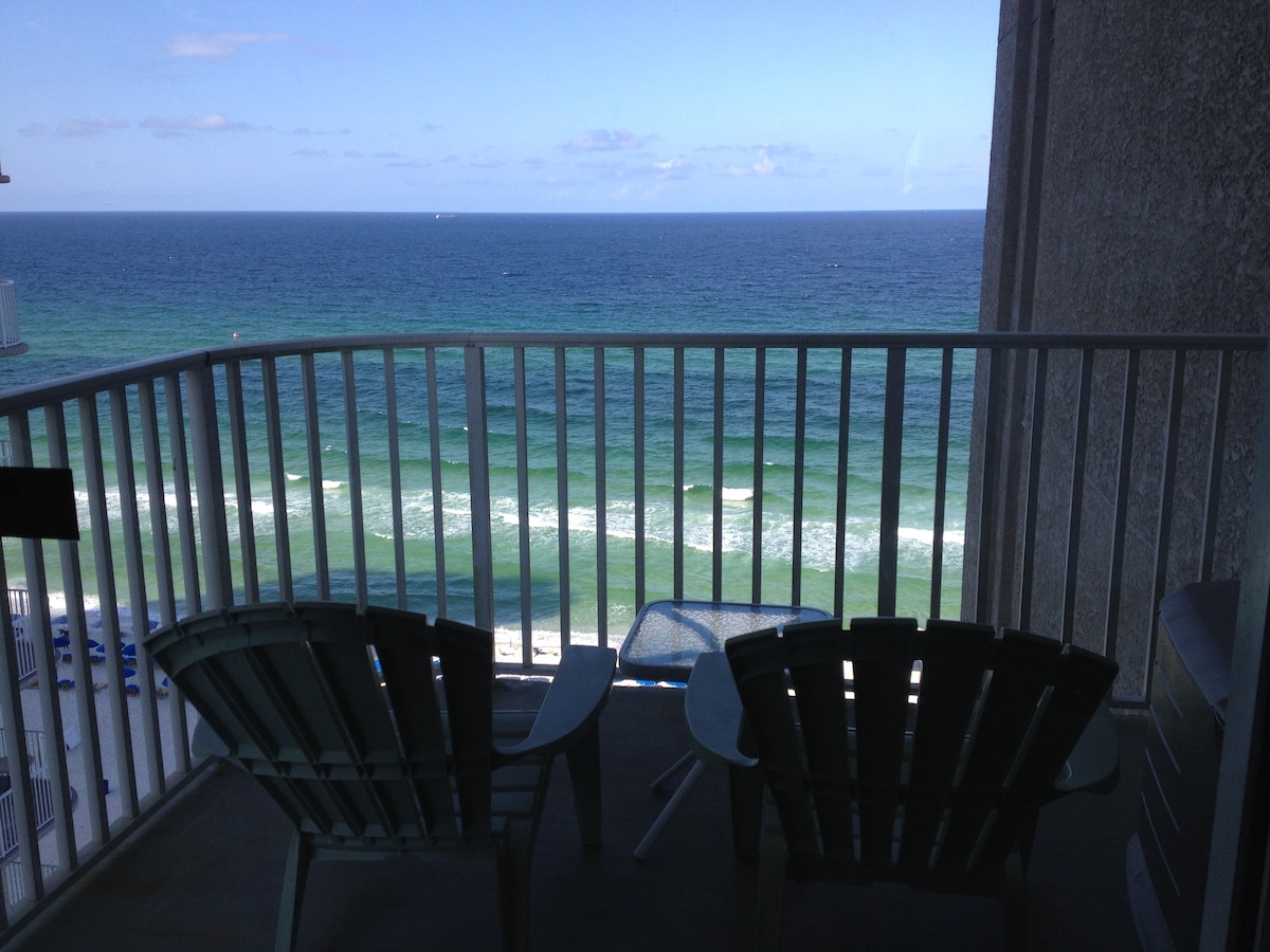 A private balcony overlooks the ocean, featuring two comfortable chairs and a small table. The view includes gentle waves and a clear blue sky, creating a serene outdoor space. The railing provides safety while enjoying the coastal scenery.