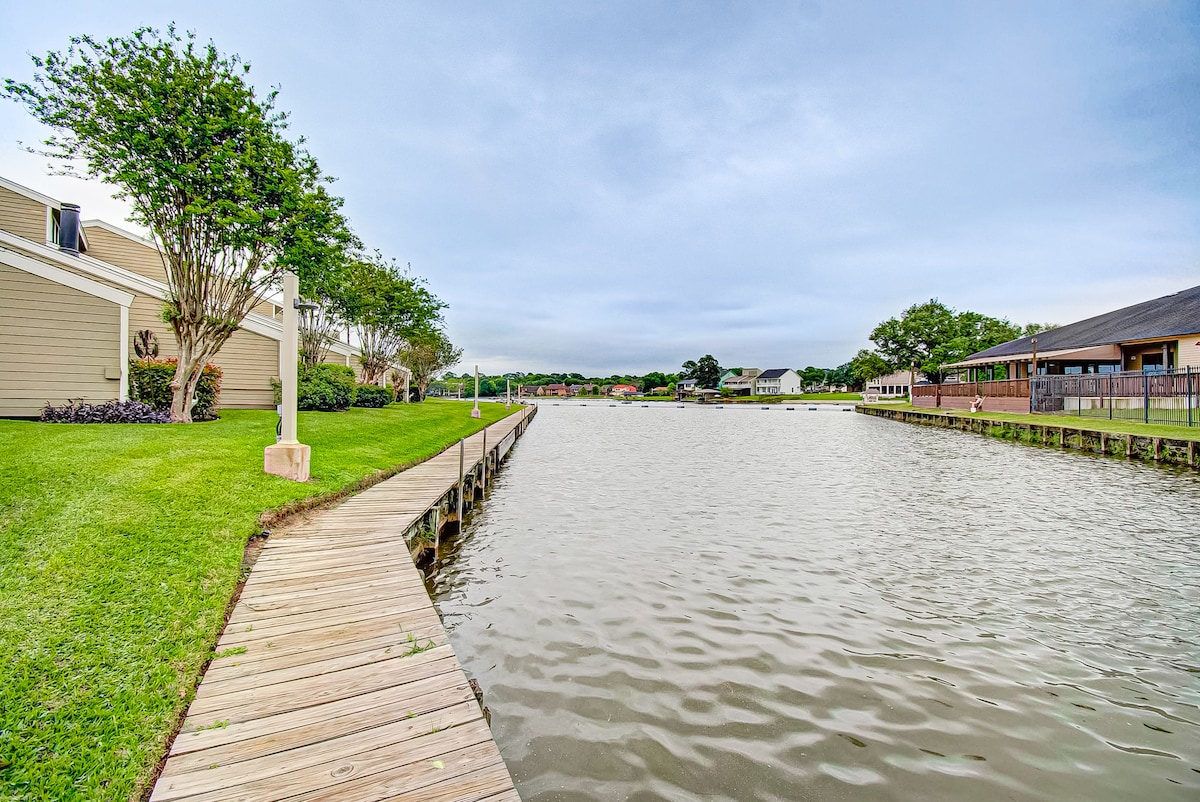A broad, wooden boardwalk extends along the waterfront, bordered by manicured grass and trees. Calm waters reflect the cloudy sky, with residential buildings visible in the distance, creating a serene lakeside environment.
