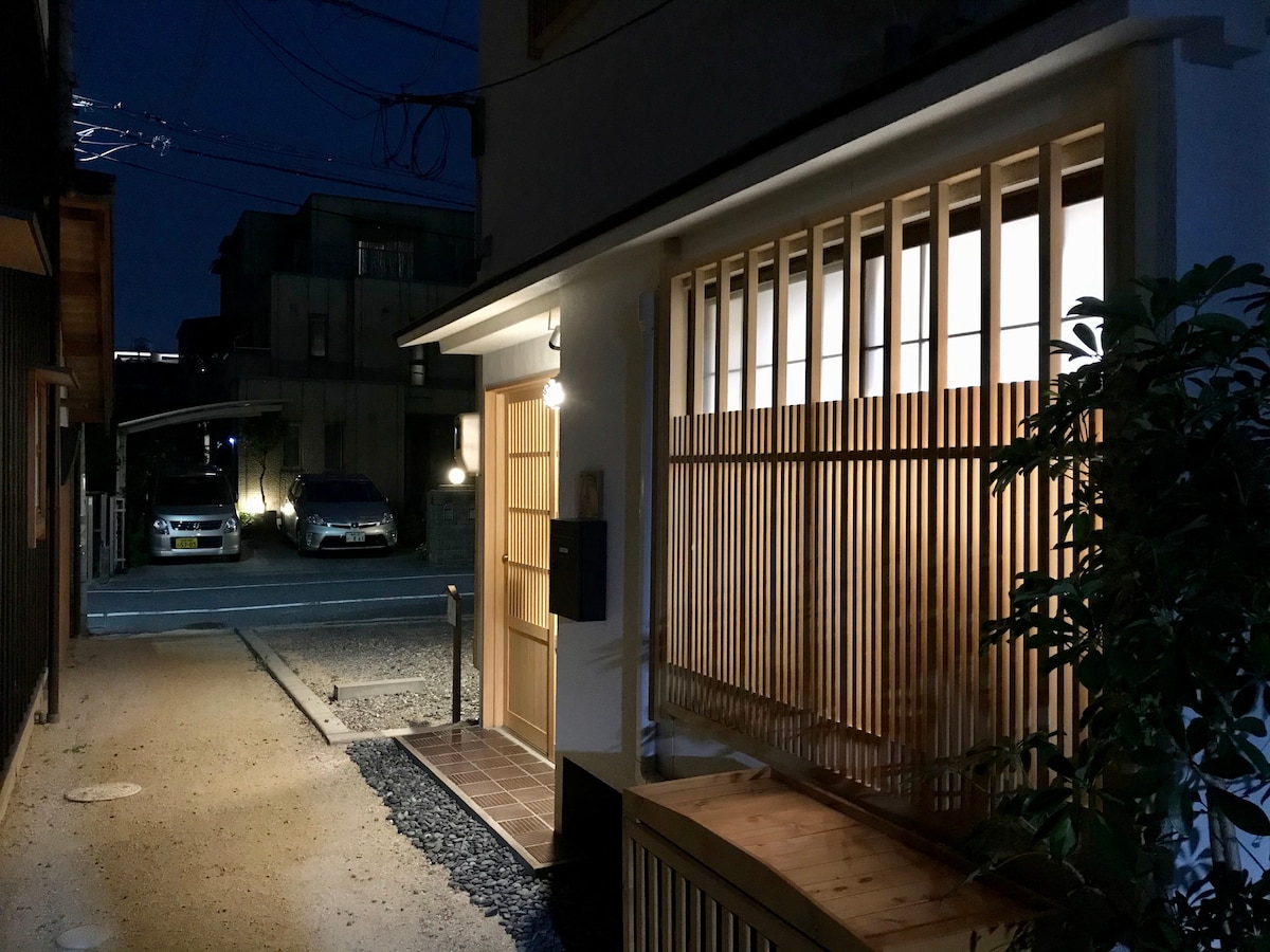 The entrance to the home features wooden slatted screens that softly illuminate the path, providing a welcoming ambiance. A tidy tiled area leads to the door, while a small mailbox is positioned nearby. The surrounding space includes a gravel pathway and glimpses of neighboring structures.