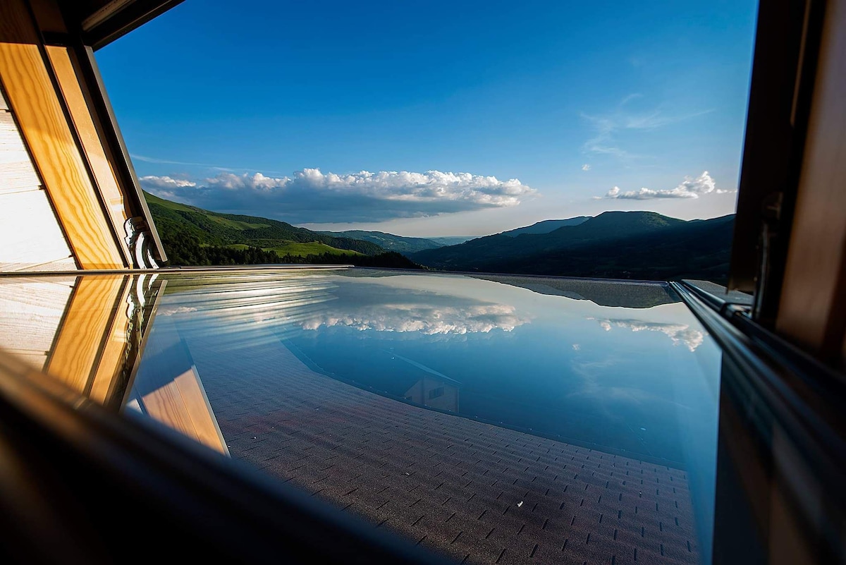 A large window frames a serene view of the mountains, reflecting the sky above. The peaceful landscape includes rolling hills and distant peaks, with clouds casting soft shadows across the terrain. The scene emphasizes the tranquility of the surrounding nature.