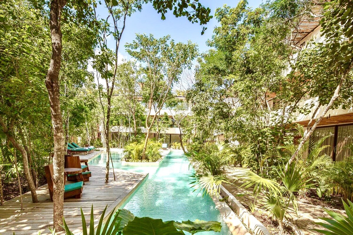 A serene jungle pool area is surrounded by lush greenery and wooden walkways. Comfortable lounge chairs are positioned nearby, providing a relaxing spot to enjoy the natural surroundings. The pool features clear waters, inviting guests to unwind in this tranquil setting.