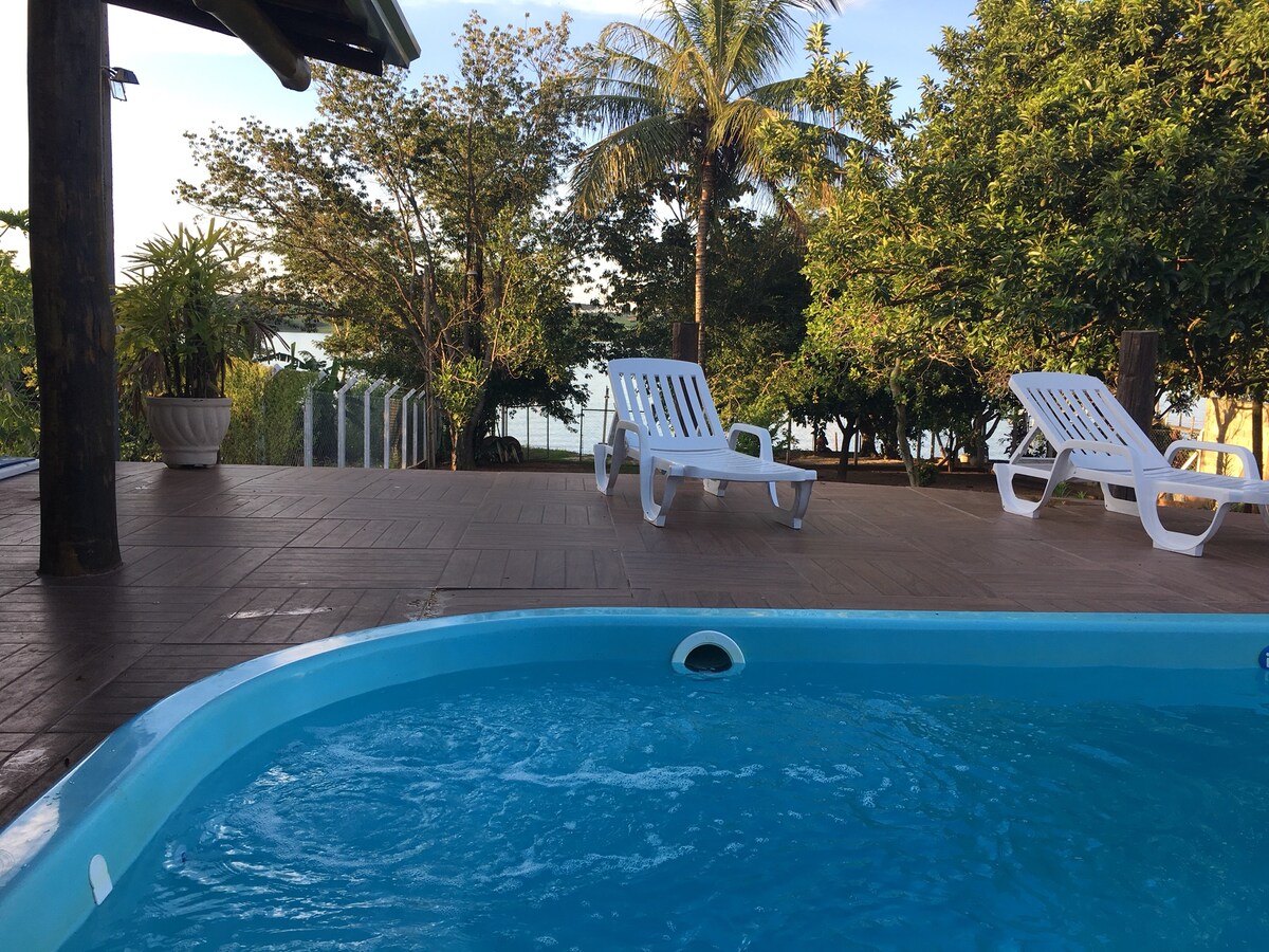 A serene outdoor area features a swimming pool with a clear blue edge, surrounded by a spacious deck. Several lounge chairs are positioned nearby, allowing for relaxation. Lush greenery and trees frame the backdrop, providing a natural setting with a view of the water.