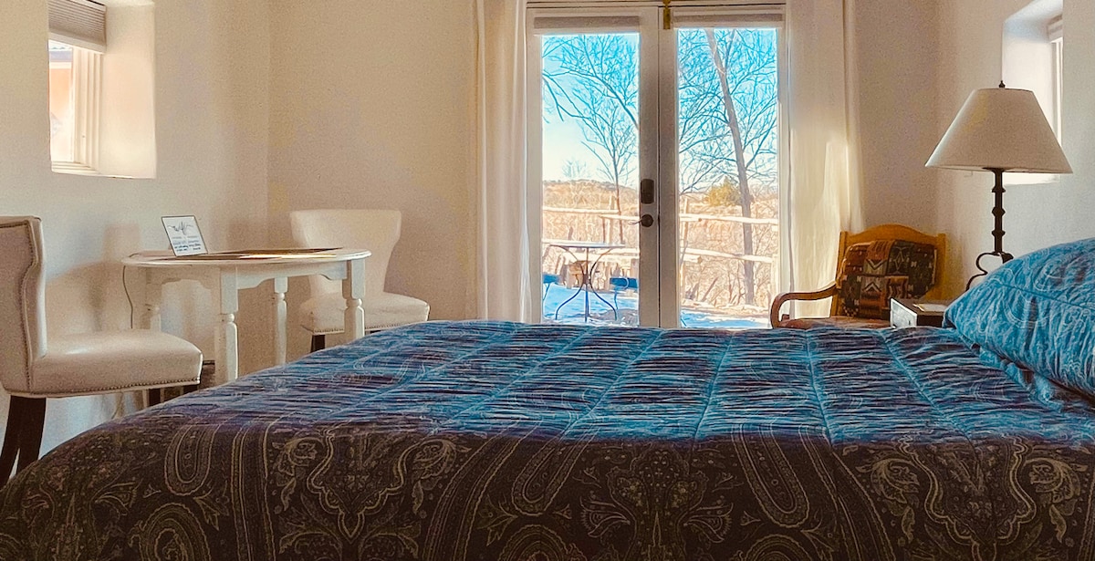 A serene sleeping area features a spacious bed covered with a patterned blanket. A small table and two chairs are positioned nearby, with natural light streaming through sliding glass doors that lead to a private patio, framed by a peaceful landscape.