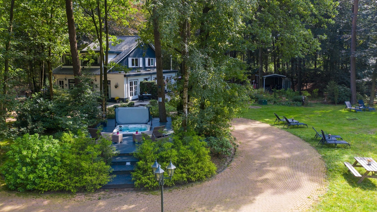 The villa is set in a lush wooded area, surrounded by greenery. A jacuzzi is positioned on a wooden deck, with pathways leading through the garden. Comfortable loungers and picnic tables are scattered throughout the expansive grassy yard, enhancing outdoor leisure possibilities.