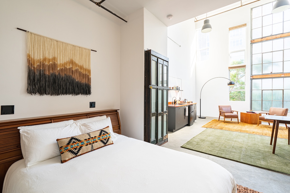 A spacious room showcases a comfortable bed with decorative pillows. Natural light illuminates the living area with large windows. A simple dining table, two chairs, and a kitchenette area are visible. A textured wall hanging adds a touch of detail above the bed.