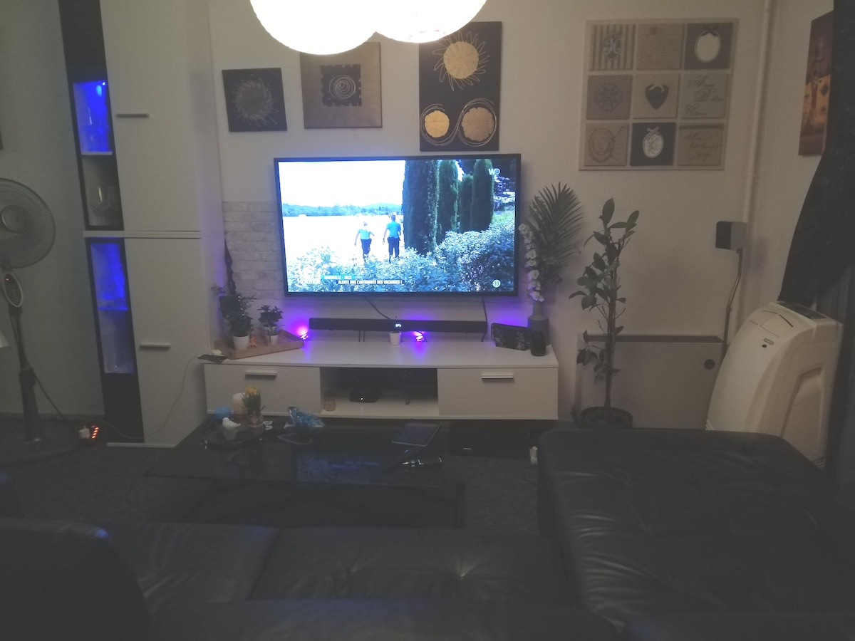 A modern living area is displayed, featuring a large television mounted on a white entertainment unit. Comfortable black leather seating is arranged around a glass coffee table. Decorative plants and wall art enhance the welcoming space, illuminated by soft lighting from a pendant fixture.
