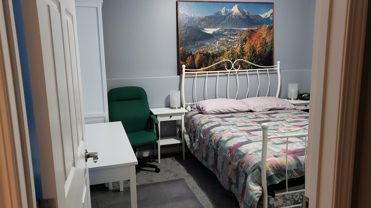 A bedroom is depicted, featuring a spacious bed adorned with a patterned quilt. A large landscape painting hangs above the bed. A writing desk and a green chair are situated beside the bed. Soft carpet covers the floor, while light colors create a calm ambiance.