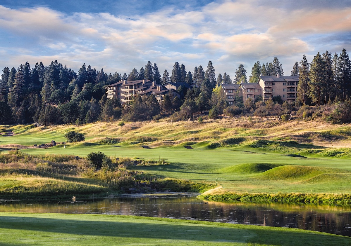 A serene view features lush green grasses of a golf course, framed by tall trees. The buildings of the resort are positioned on a gentle ridge in the background, overlooking a calm water body reflecting the surroundings.
