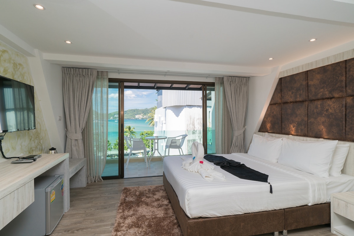 A spacious room features a queen size bed and a single bed, with large glass doors opening to a private balcony offering a sea view. Soft light floods the space, and a TV is mounted on the wall. A light-colored desk and comfortable seating area are also visible.