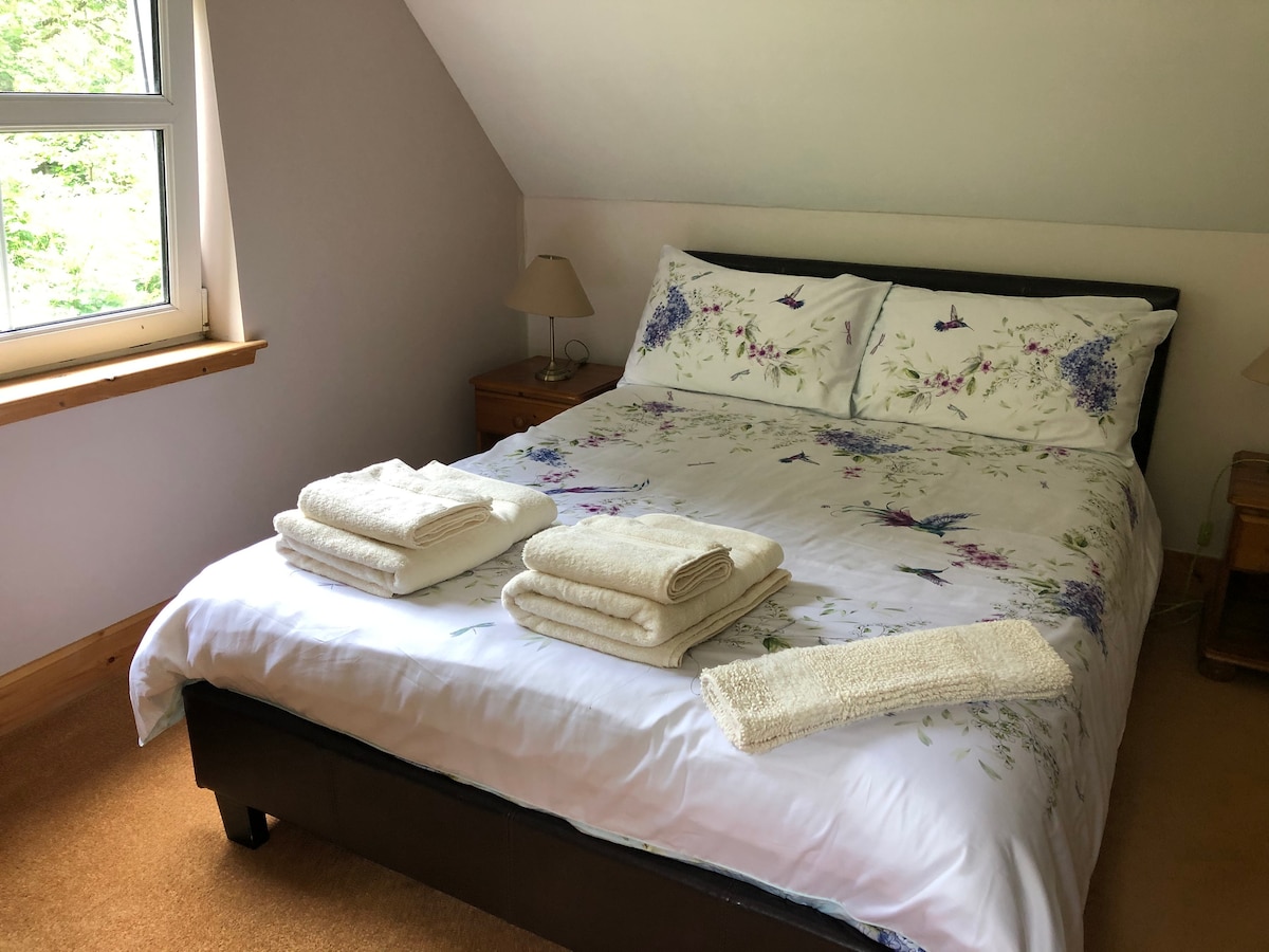 A serene bedroom is presented with a spacious bed adorned in floral-patterned bedding. Freshly laundered towels are neatly arranged at the foot of the bed, and soft bedside lamps are placed on wooden tables. Natural light filters through a large window, illuminating the room.