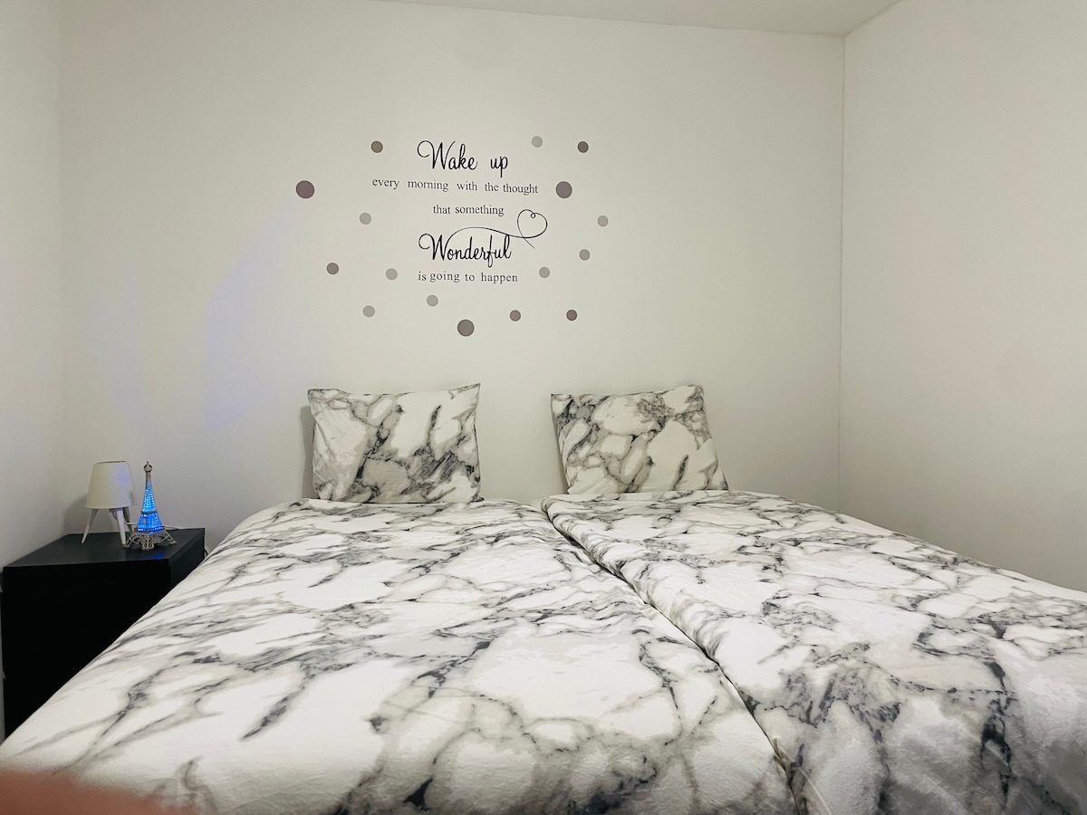 A cozy sleeping area features a marble-patterned bedding spread across two separate beds. A nightstand with a small lamp is positioned beside the beds. Inspirational wall art is displayed above, complementing the simple, welcoming design of the room.