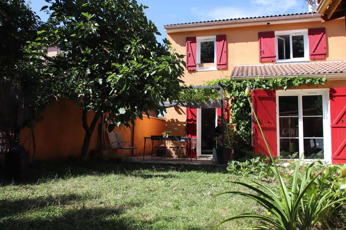 The exterior of the house features a vibrant yellow façade with red shutters. A small garden is seen with green grass and shrubbery. A patio area is present, furnished with a table and chairs, partially shaded by a nearby tree.