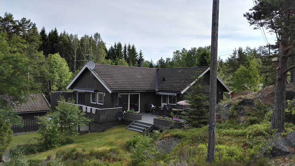 A spacious house is nestled among greenery, surrounded by trees and shrubs. The structure features a dark exterior with large windows and a deck area. A terrace is visible, suggesting outdoor seating. Rocky terrain and well-maintained grass add to the natural setting.