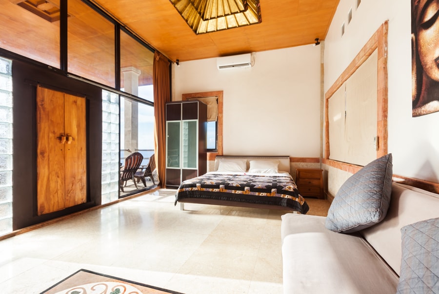 A spacious room is illuminated by natural light, showcasing a cozy bed adorned with a decorative blanket. A comfortable sofa sits nearby, and large glass doors provide a view of the outside. Artwork and a glass wardrobe add visual interest to the serene space.
