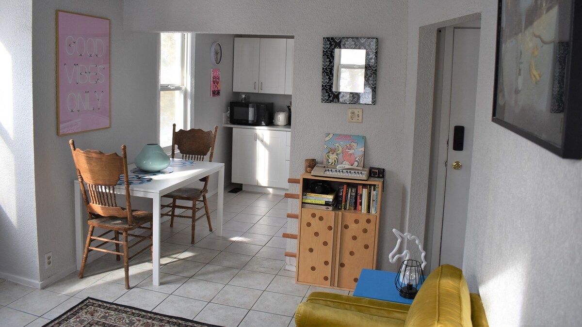 A bright living space showcases a dining area with two wooden chairs beside a white table. The kitchen is visible in the background, equipped with modern appliances. A yellow sofa and a bookshelf add a pop of color, with light entering through large windows.